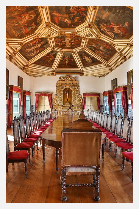 Museu da Misericórdia - Vista do Salão Nobre do Museu com mesa grande e mais de vinte cadeiras de madeira ao redor, teto com telas pintadas a mão e um cristo na cruz ao fundo